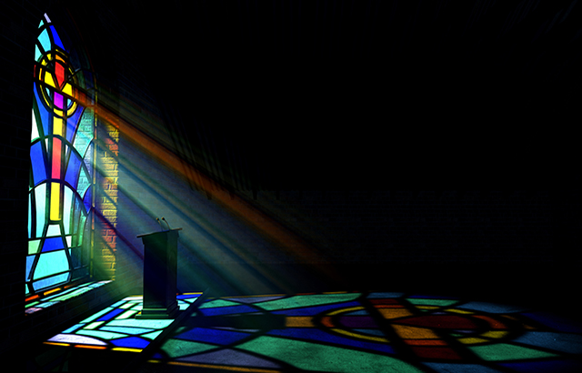 A dim old church interior lit by suns rays penetrating through a colorful stained glass window in the pattern of a crucifix reflecting colours on the floor and a speech pulpit