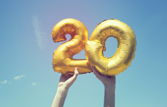 A gold foil number 20 balloon is held high in the air by caucasian male hand. The image has been taken outdoors on a bright sunny day, the sky is blue with some clouds. A vintage style effects has been added to the image.