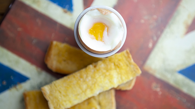 A broken boiled egg in an egg cup with buttered toast soldiers on a British flag surface viewed from a high angle with shallow depth of field