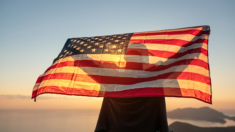 Proud man raising the flag of the United States of America standing on the top of the mountain at sunset