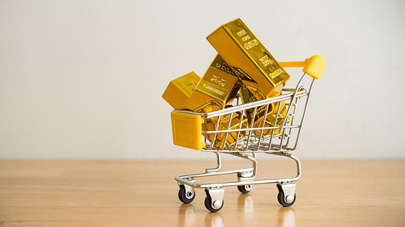 Gold bars in shopping trolley on wooden table with white wall background copy space. Gold price rising all time high, high demand purchasing, investment in commodity market, etf, future concept.